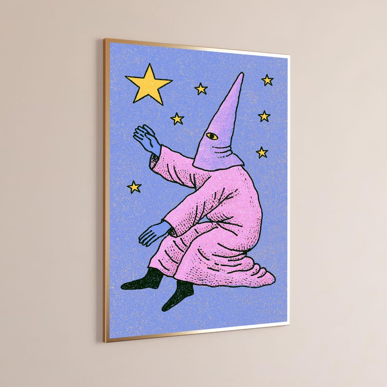 May include: A framed wall art print featuring a figure in a pink hooded robe with a single yellow eye on the hood. The figure has blue hands and black feet, set against a textured periwinkle blue background with several yellow stars of varying sizes.