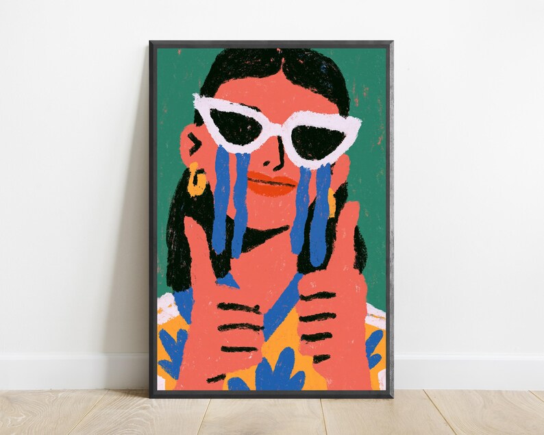 May include: A framed art print featuring a stylized illustration of a person with dark hair wearing white cat-eye sunglasses. The person has blue tears streaming down their face, is giving a double thumbs-up, and wears a yellow and blue patterned shirt.