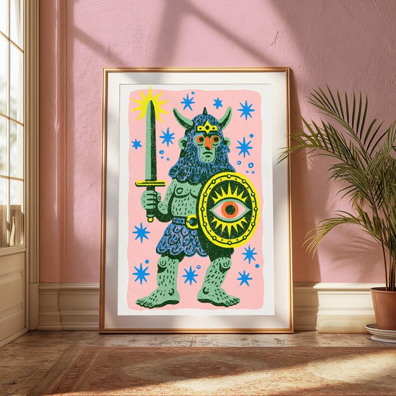 May include: A framed art print featuring a green, horned, bearded warrior figure holding a sword and a yellow shield with a central eye motif. The character is set against a light pink background decorated with scattered blue star shapes.