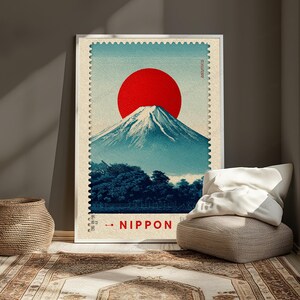 May include: A rectangular wall art print designed to resemble a postage stamp, featuring a large red sun positioned above a snow-capped mountain peak. The base of the mountain is surrounded by dark blue trees, and the text NIPPON is printed in red.