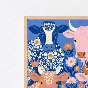May include: A framed wall art print featuring a blue cow with a white floral pattern, a pink cow, and a smaller peach-coloured cow. The cows are surrounded by stylised orange and white flowers against a solid blue background in a light wood frame.