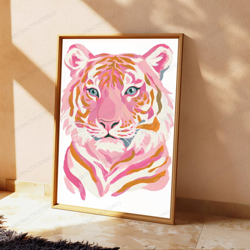 May include: A framed wall art print featuring a stylized tiger portrait. The tiger is rendered in shades of pink, white, and orange with distinct stripes and light blue eyes. The artwork is contained within a simple, light-colored wooden frame.