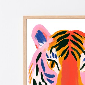 May include: A framed art print featuring a stylized tiger head illustration. The tiger is rendered in vibrant colors including pink, bright orange, and deep red, with bold dark green stripes and blue accents on the ears and face, set in a light wood frame.
