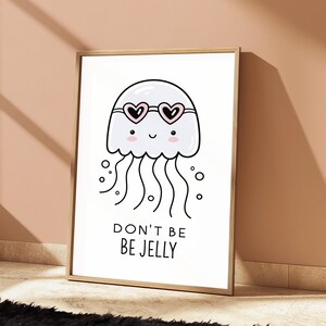 May include: A framed wall art print featuring a white cartoon jellyfish with pink cheeks and heart-shaped sunglasses. The jellyfish has black outlines and wavy tentacles. Below the illustration, the text reads DON'T BE BE JELLY in black letters.
