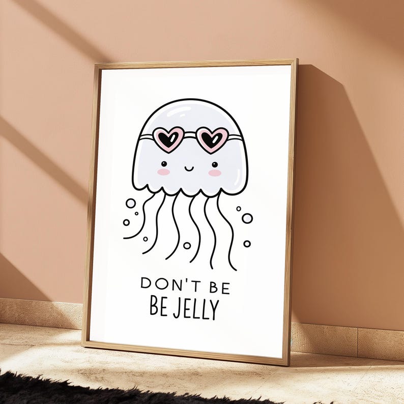 May include: A framed wall art print featuring a white cartoon jellyfish with pink cheeks and heart-shaped sunglasses. The jellyfish has black outlines and wavy tentacles. Below the illustration, the text reads DON'T BE BE JELLY in black letters.