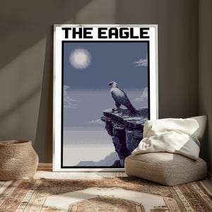 May include: A framed wall art print featuring a pixelated illustration of an eagle perched on a rocky cliff under a full moon. The text THE EAGLE is printed in bold black block letters at the top of the white-bordered rectangular poster.