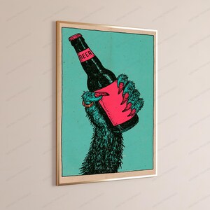 May include: A framed wall art print featuring a dark, furry creature hand with sharp claws holding a black beer bottle. The bottle has a pink label with the word BEER printed in black. The background is a solid teal colour with a black border.