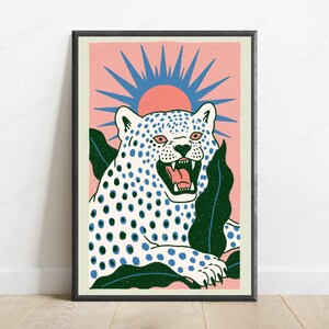 May include: A framed art print featuring a white leopard with blue spots and an open mouth, set against a pink background with a blue sunburst graphic. The leopard is surrounded by dark green tropical leaves, all enclosed in a black frame.