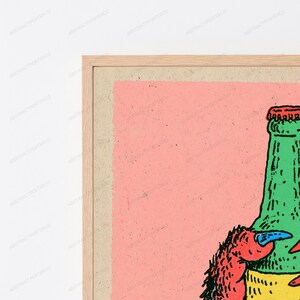 May include: A framed art print featuring a graphic illustration of a green bottle with a red cap and a red creature with a blue claw. The artwork is set against a solid light pink background, enclosed within a light-toned wooden frame.