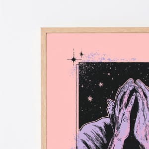 May include: A framed art print featuring a light pink border and a central black rectangular panel filled with white stars. The illustration depicts two lavender-colored hands held together in a gesture against the starry night sky background.