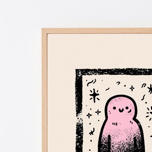 May include: A framed print featuring a pink, rounded character with a simple smiling face, surrounded by black ink-style stars and squiggles. The artwork is enclosed in a light-colored wooden frame, set against a plain, off-white background.