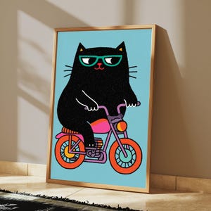 K&ouml;nnte beinhalten: Ein gerahmter Kunstdruck zeigt eine schwarze Katze mit t&uuml;rkisfarbener Brille auf einem rosa-violetten Motorrad mit orangefarbenen R&auml;dern vor hellblauem Hintergrund. Die Illustration verwendet kr&auml;ftige Linien und fl&auml;chige Farben f&uuml;r das Motiv.