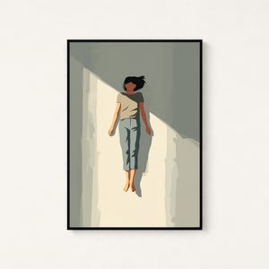 May include: A framed wall art print featuring a stylized illustration of a person with dark hair wearing a light brown t-shirt and blue pants, floating in a vertical shaft of light against a muted gray background with sharp shadows.
