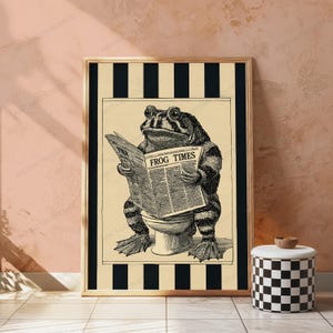 May include: A framed wall art print featuring a black and white illustration of a toad wearing glasses while sitting on a toilet and reading a newspaper titled THE FROG TIMES. The background consists of vertical black and white stripes.