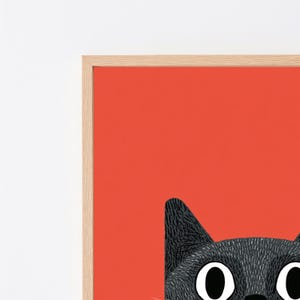 May include: A framed wall art print featuring a stylized black cat with large white eyes and black pupils set against a solid vibrant orange background. The artwork is enclosed in a light-toned wooden frame with a clean, simple border design.