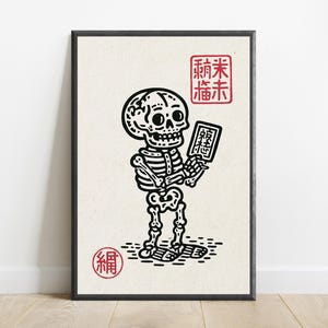 May include: A framed art print featuring a black ink illustration of a skeleton holding a small rectangular tablet. The design includes red square and circular stamps with Asian characters, set against a light cream-colored background.