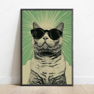 May include: A framed vertical wall art print featuring a detailed illustration of a cat wearing large black sunglasses. The cat is centered against a sage green background with radiating light lines emanating from behind its head in a sunburst pattern.