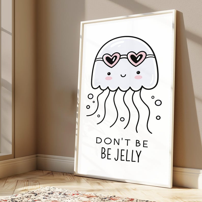 May include: A framed white wall art print featuring a cartoon jellyfish wearing pink heart-shaped sunglasses. The jellyfish has a smiling face with pink cheeks and wavy tentacles. Below the illustration, the text reads: DON'T BE BE JELLY.