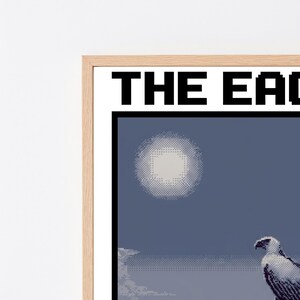May include: A framed pixel art print featuring a white eagle perched on a dark blue background with a pale circular moon. The text THE EAGLE is printed in bold black capital letters at the top of the white border within a light wood frame.