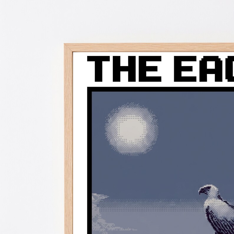 May include: A framed pixel art print featuring a white eagle perched on a dark blue background with a pale circular moon. The text THE EAGLE is printed in bold black capital letters at the top of the white border within a light wood frame.
