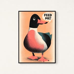 May include: A framed wall art print featuring a stylised illustration of a duck with a black head, a coral-pink chest, and dark teal wings. The duck has bright orange webbed feet and a speech bubble above its head containing the text FEED ME!