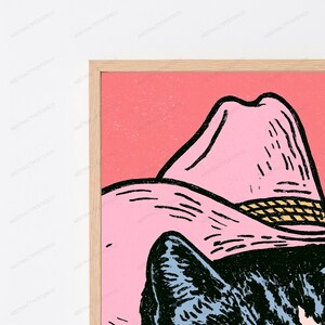 May include: A framed wall art print featuring a black cat wearing a light pink cowboy hat with a yellow band, set against a solid coral pink background. The illustration uses bold black outlines and a textured, graphic style within a light wood frame.