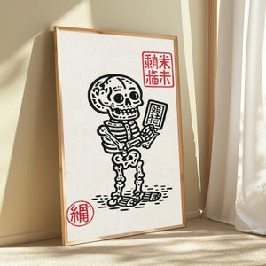May include: A framed wall art print featuring a black line drawing of a skeleton holding a small rectangular sign. The print includes red decorative stamps with Asian-style characters in the top right and bottom left corners on a cream background.