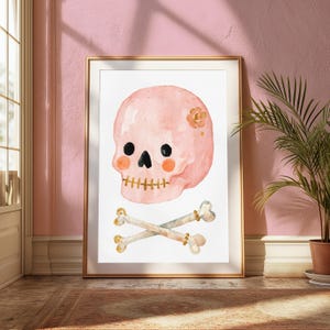 Puede incluir: Una l&aacute;mina de acuarela enmarcada que muestra una calavera de color rosa claro con ojos negros, nariz negra y manchas naranjas en las mejillas. La calavera tiene una boca dorada y una peque&ntilde;a flor dorada en el lateral. Debajo hay dos huesos cruzados.
