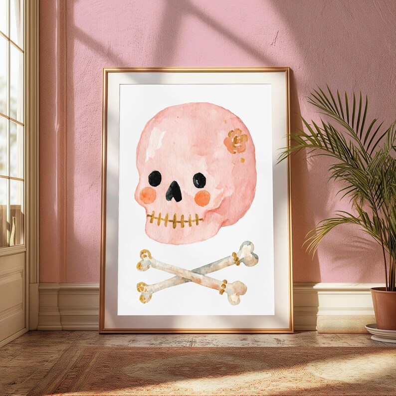 Puede incluir: Una l&aacute;mina de acuarela enmarcada que muestra una calavera de color rosa claro con ojos negros, nariz negra y manchas naranjas en las mejillas. La calavera tiene una boca dorada y una peque&ntilde;a flor dorada en el lateral. Debajo hay dos huesos cruzados.