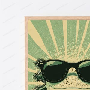May include: A framed wall art print featuring a close-up illustration of an axolotl wearing black sunglasses. The background displays a light green and cream sunburst pattern with a distressed, textured effect. The frame is light-colored wood.