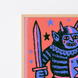 May include: A framed art print featuring a horned creature in a striped shirt holding a sword and shield. The illustration uses a vibrant color palette of coral pink, deep blue, and black, with small star and circle motifs scattered across the background.