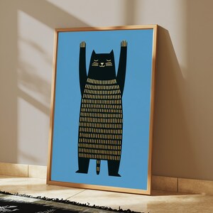 Op de afbeelding: Een ingelijste kunstprint met een gestileerde zwarte kat met de voorpoten omhoog tegen een effen lichtblauwe achtergrond. Het lichaam van de kat heeft een textuurpatroon van verticale gouden lijnen en de ogen zijn gesloten in een dunne gouden lijst.