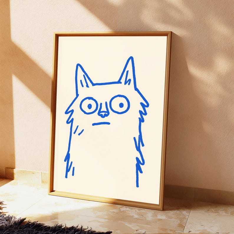 May include: A framed wall art print featuring a simple, bold blue line drawing of a cat with large, wide eyes and a fluffy, jagged outline. The illustration is centered on a plain, light cream-colored background within a light wood frame.