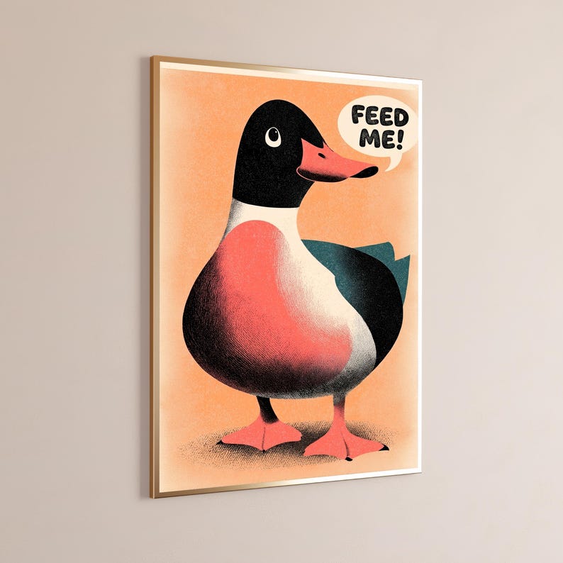 May include: A framed wall art print featuring a stylised illustration of a duck with a black head, a pink chest, and dark teal wings against a light orange background. A white speech bubble above the duck contains the black text FEED ME!