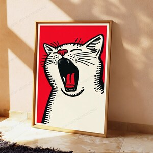 May include: A framed wall art print featuring a stylized white cat with black line details and a red nose, shown with its mouth wide open in a yawn. The cat is set against a solid red background, and the entire image is enclosed in a light wood frame.