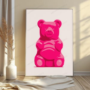 May include: A framed wall art print featuring a centered, vibrant hot pink gummy bear illustration against a solid, light cream-colored background. The bear is depicted in a simple, graphic style with rounded features and a smooth, glossy appearance.