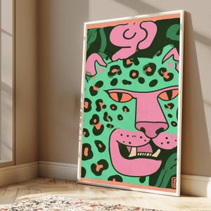 May include: A framed rectangular wall art print featuring a stylized leopard illustration. The leopard has a bright mint green face with black spots, a pink snout, and orange eyes. The background includes dark green foliage and pink abstract shapes.