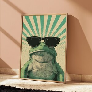 K&ouml;nnte beinhalten: Ein gerahmter Kunstdruck, der einen gr&uuml;nen Frosch mit schwarzer Sonnenbrille zeigt. Der Hintergrund besteht aus einem hellbeigen Feld mit strahlenden t&uuml;rkisfarbenen Sonnenstrahlen und einem dezenten Punktmuster, eingefasst in einen Holzrahmen.