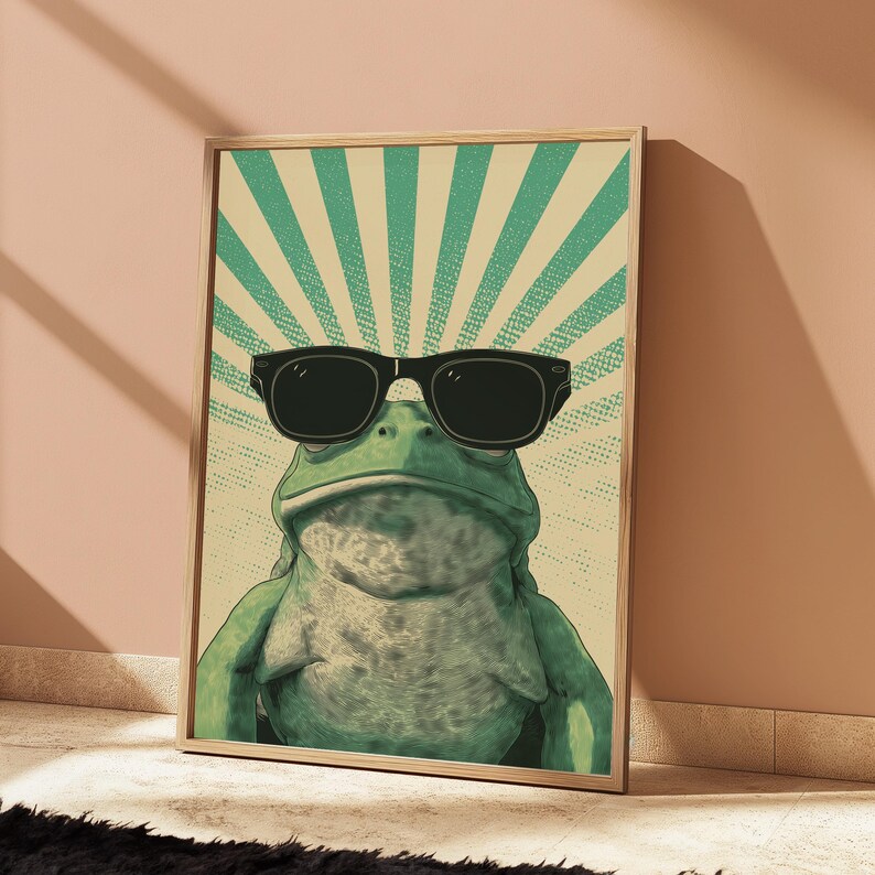 K&ouml;nnte beinhalten: Ein gerahmter Kunstdruck, der einen gr&uuml;nen Frosch mit schwarzer Sonnenbrille zeigt. Der Hintergrund besteht aus einem hellbeigen Feld mit strahlenden t&uuml;rkisfarbenen Sonnenstrahlen und einem dezenten Punktmuster, eingefasst in einen Holzrahmen.