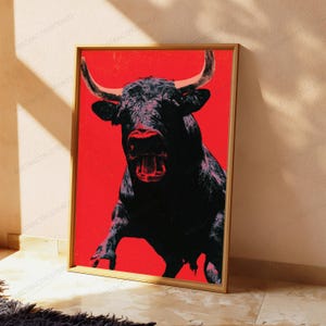 K&ouml;nnte beinhalten: Ein rechteckiger gerahmter Kunstdruck mit einer kontrastreichen, stilisierten Illustration eines schwarzen Stiers mit markanten H&ouml;rnern vor einem einfarbigen, leuchtend roten Hintergrund. Der Stier ist frontal mit offenem Maul dargestellt.