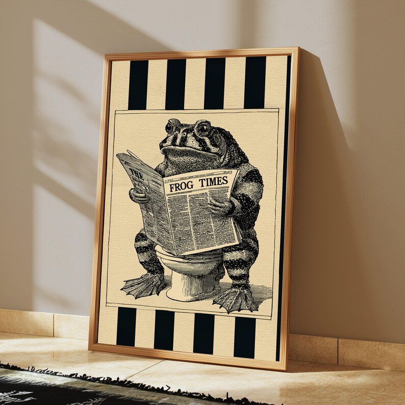 May include: A framed wall art print featuring a black and white illustration of a toad sitting on a toilet reading a newspaper titled The Frog Times. The background consists of vertical black and beige stripes, and the toad has textured skin.