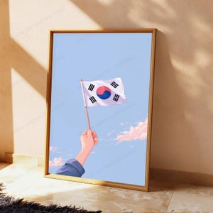 May include: A framed vertical art print featuring a hand holding a small South Korean flag against a light blue sky with soft pink clouds. The flag displays the central red and blue taegeuk symbol surrounded by four black trigrams on a white field.