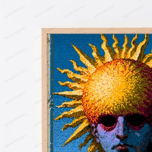 May include: A framed pixel art print featuring a stylized sun with golden rays radiating from a central circular form. Below the sun, a partial human face is depicted in shades of blue and purple with dark, hollow eye sockets and a neutral expression.