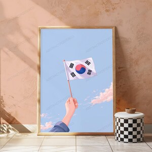 May include: A framed vertical poster featuring an illustration of a hand holding a South Korean flag against a light blue sky with soft pink clouds. The flag displays the central red and blue taegeuk symbol surrounded by four black trigrams.