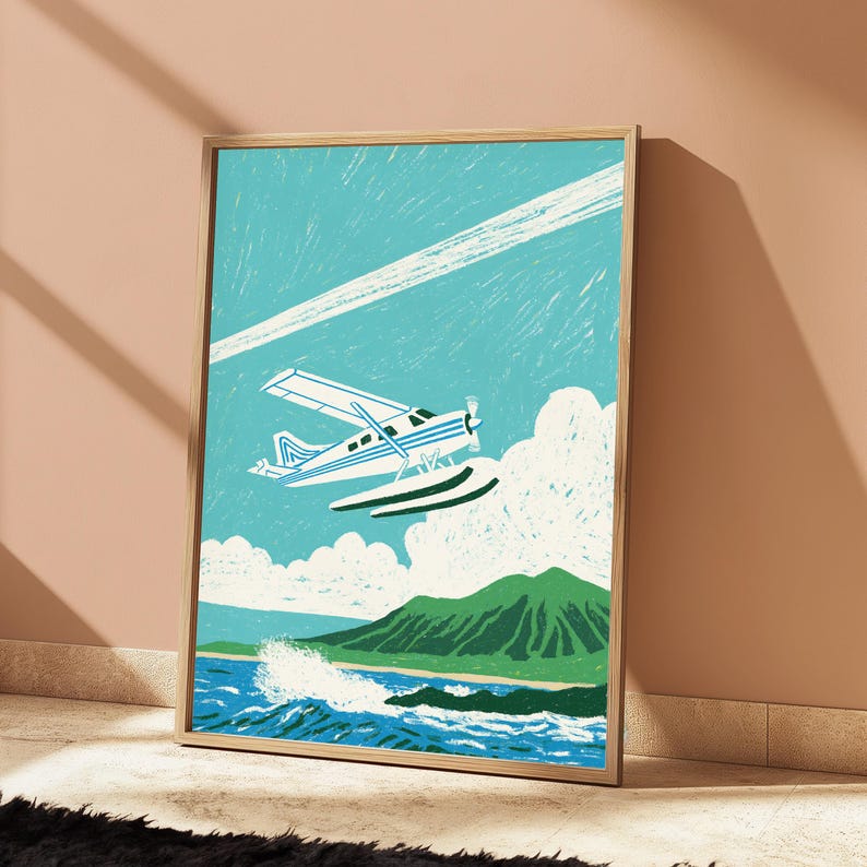 May include: A framed art print featuring a stylized illustration of a white and blue seaplane flying over a green mountain and blue ocean waves. The sky is a bright turquoise with a white contrail and fluffy white clouds in the background.