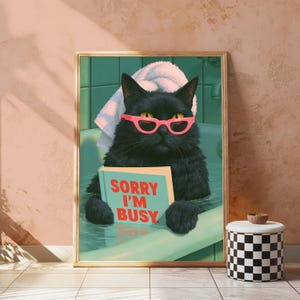 May include: A framed wall art print featuring a black cat in a bathtub wearing pink cat-eye glasses and a white towel on its head. The cat holds a light blue book with the red text SORRY I'M BUSY printed on the cover. The background shows green tiles.