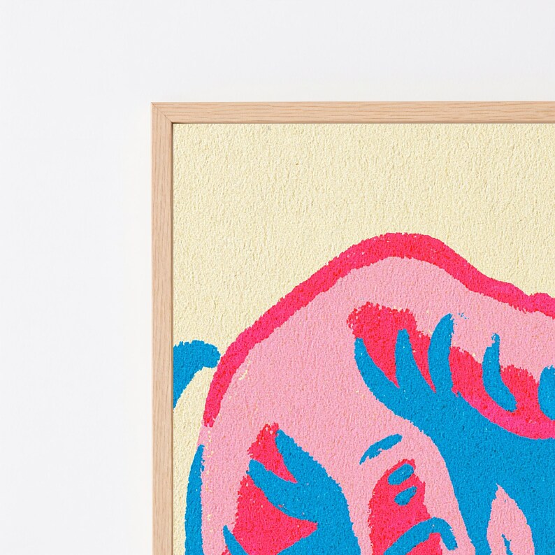 May include: A framed wall art print featuring a textured cream background with a large, abstract pink shape layered with vibrant blue, hand-drawn style markings. The artwork is enclosed in a light-toned wooden frame with a visible grain pattern.