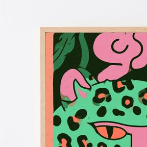 May include: A framed wall art print featuring a stylized green leopard with black spots, bright orange eyes, and pink accents against a dark background. The artwork is enclosed in a light-colored wooden frame with a thin, vertical peach-colored border.