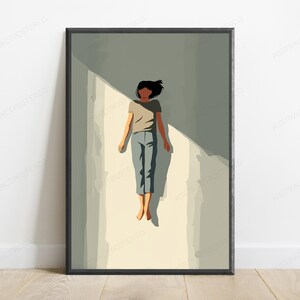 May include: A framed wall art print featuring a stylized illustration of a person with dark hair wearing a beige t-shirt and blue pants, lying flat on a light-colored surface with a sharp diagonal shadow cast across the scene in shades of gray.