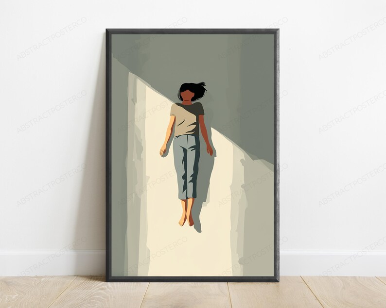 May include: A framed wall art print featuring a stylized illustration of a person with dark hair wearing a beige t-shirt and blue pants, lying flat on a light-colored surface with a sharp diagonal shadow cast across the scene in shades of gray.